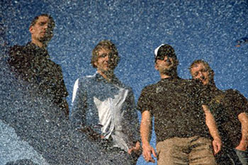 Photo de groupe pour Vancouver. Quatre des membres, reflétés dans une flaque d'eau.