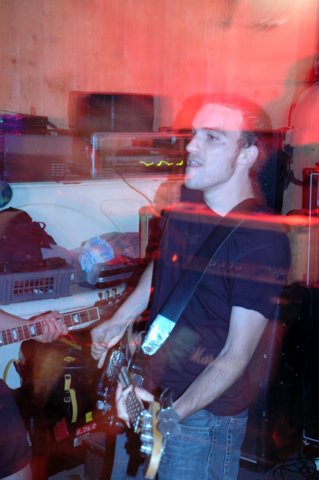 Une photo du bassiste du groupe Aghast, à travers lequel on voit tout le mobilier qui se trouve derrière lui.