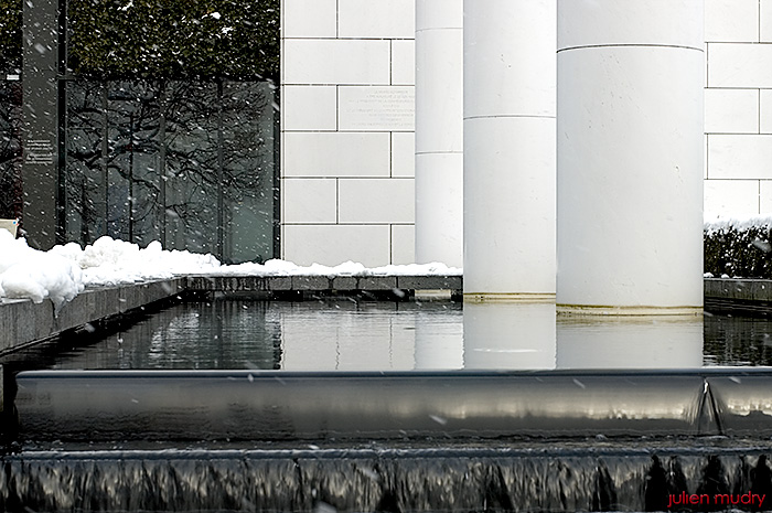 Une fontaine moderne dont sortent des colonnes, le tout sous la neige qui tombe.